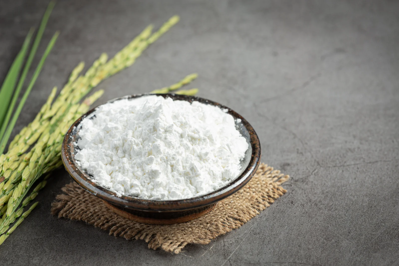 farinha-de-arroz Tigela de cerâmica escura cheia de farinha branca sobre um pedaço de juta, com espigas de arroz verde à esquerda, em um fundo cinza escuro