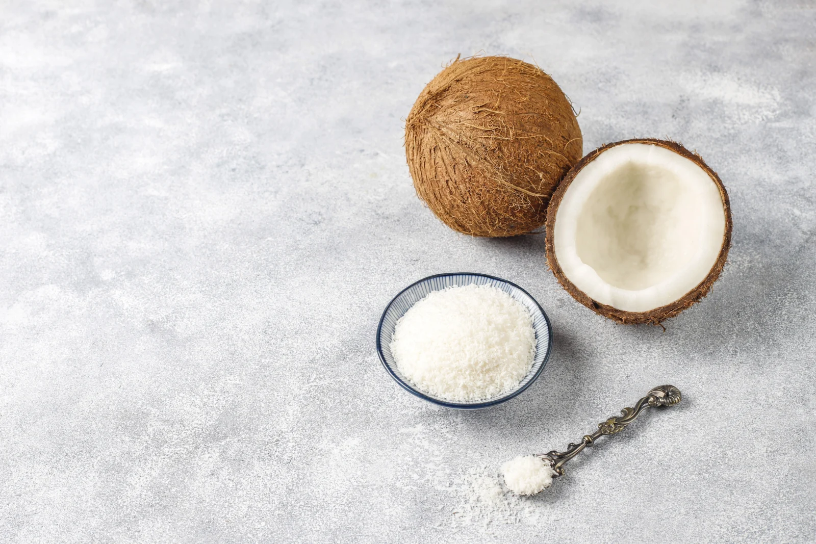 farinha-de-coco Um coco inteiro, um coco partido ao meio, uma tigela pequena com coco ralado e uma colher de metal com coco ralado, tudo sobre uma superfície cinza clara