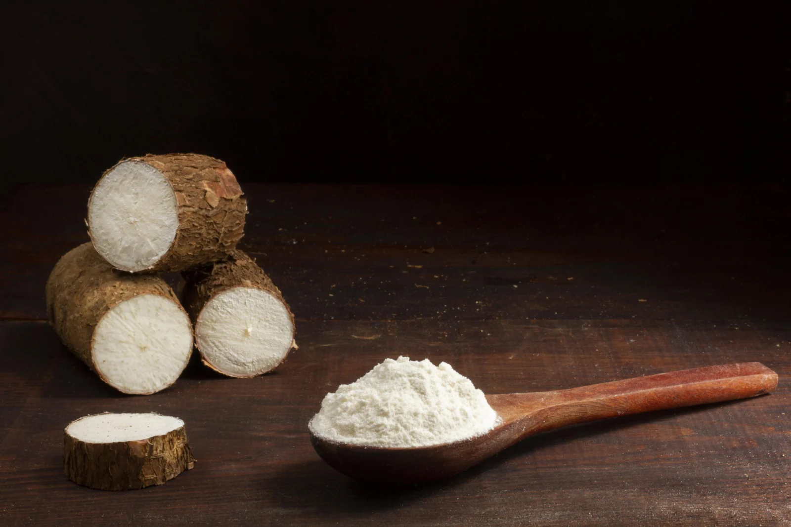 farinha-de-mandioca Três pedaços de mandioca descascados e cortados, um deles em fatia, ao lado de uma colher de pau cheia de farinha branca, sobre uma mesa de madeira escura