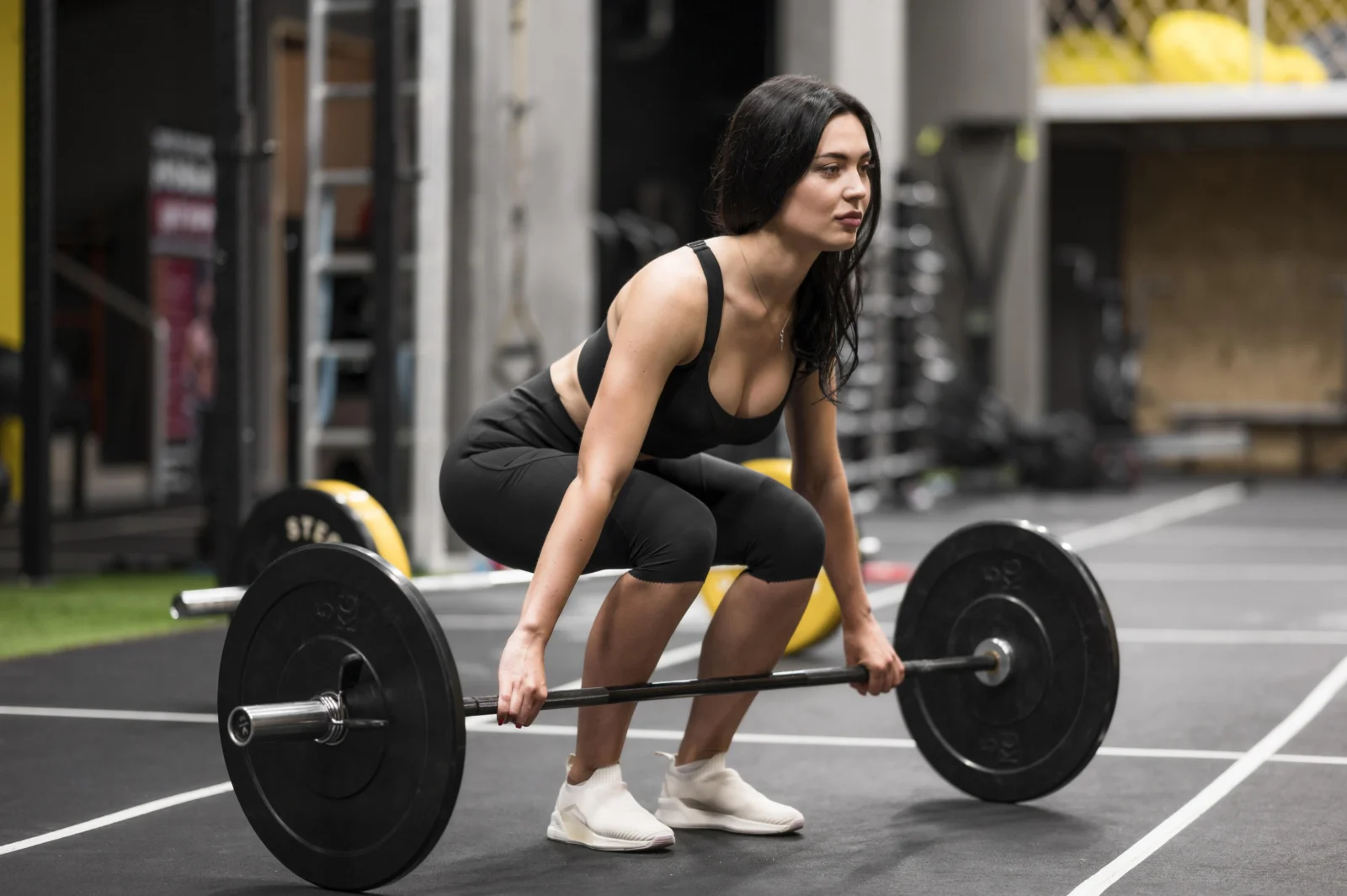 Mulher de cabelo escuro, vestindo top e calça legging pretos, agachada para levantar uma barra com pesos em uma academia. Ela tem expressão concentrada e está em posição de levantamento terra, com o olhar fixo à frente